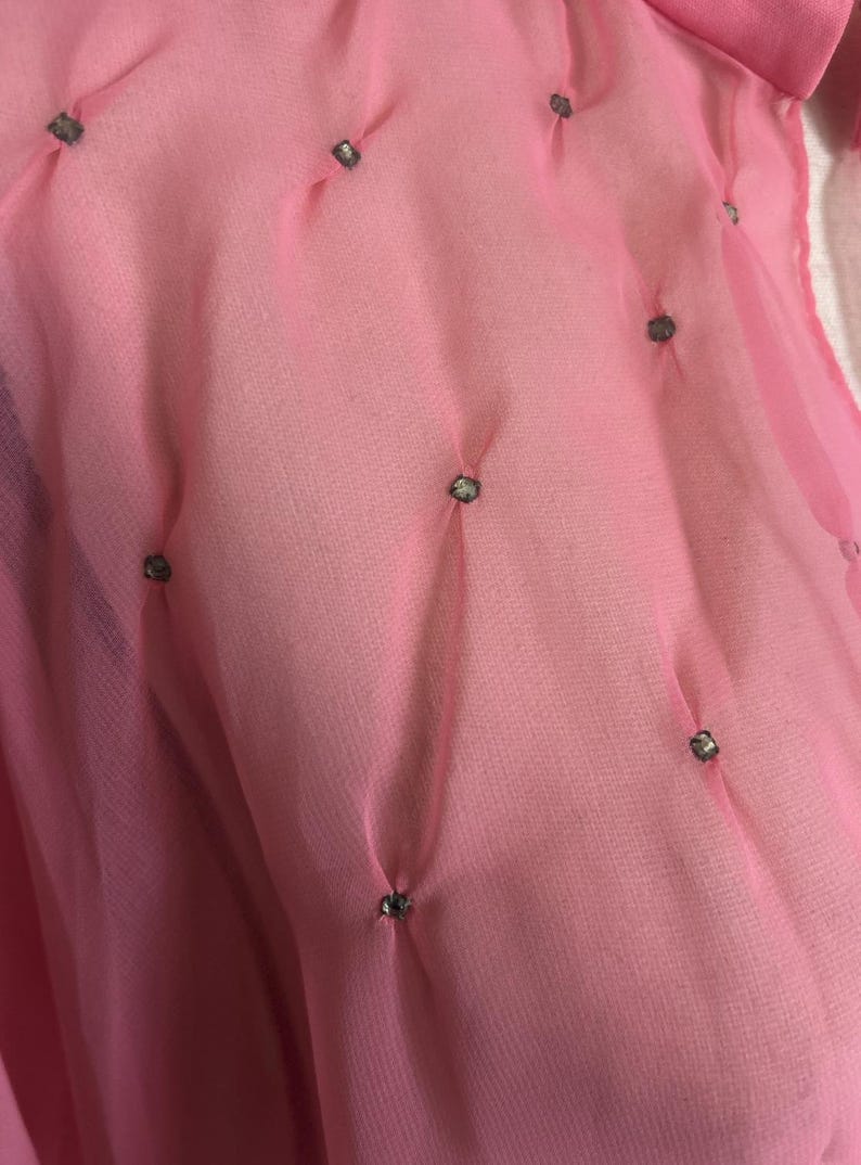 May include: Close-up of a sheer pink fabric with a textured appearance. Small, decorative embellishments are evenly spaced across the surface. The fabric is gathered at each embellishment, creating a subtle, dimensional effect.