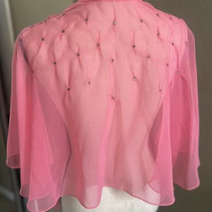 May include: A pink, sheer, cape-style top with a high collar and small, decorative embellishments. The top has long, flowing sleeves and a delicate, lightweight appearance. The fabric is a soft, pastel pink, and the design is simple and elegant.