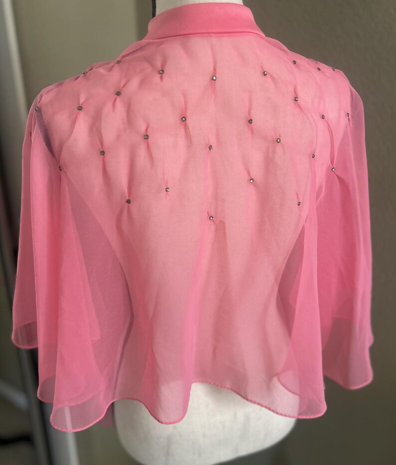 May include: A pink, sheer, cape-style top with a high collar and small, decorative embellishments. The top has long, flowing sleeves and a delicate, lightweight appearance. The fabric is a soft, pastel pink, and the design is simple and elegant.