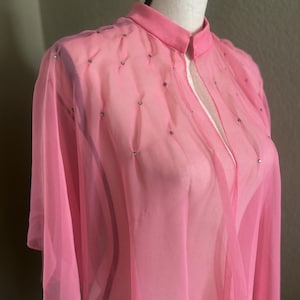 May include: A sheer, light pink cape-style garment with a mandarin collar and short sleeves. The fabric is embellished with small, silver-colored accents. The cape is displayed on a mannequin, showcasing its flowing design and delicate texture.