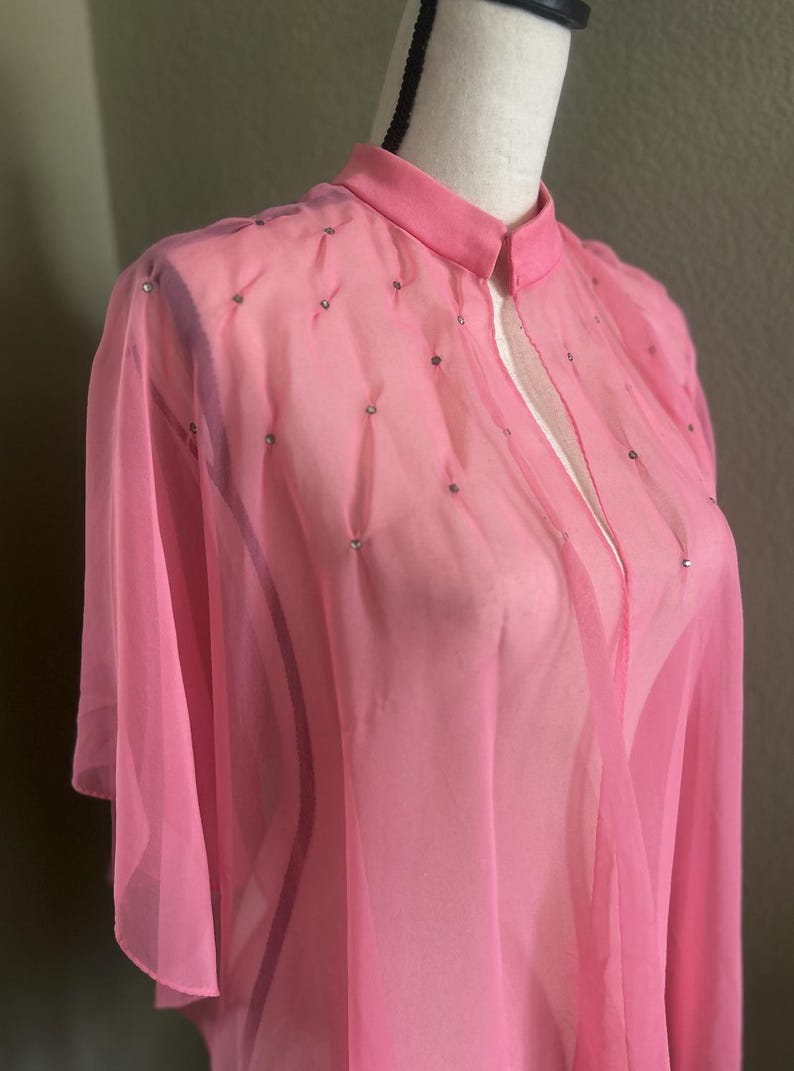 May include: A sheer, light pink cape-style garment with a mandarin collar and short sleeves. The fabric is embellished with small, silver-colored accents. The cape is displayed on a mannequin, showcasing its flowing design and delicate texture.