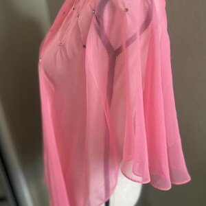 May include: A sheer, light pink top with a high collar and flared sleeves. The fabric is embellished with small, scattered silver-colored rhinestones. The garment is displayed on a white mannequin.