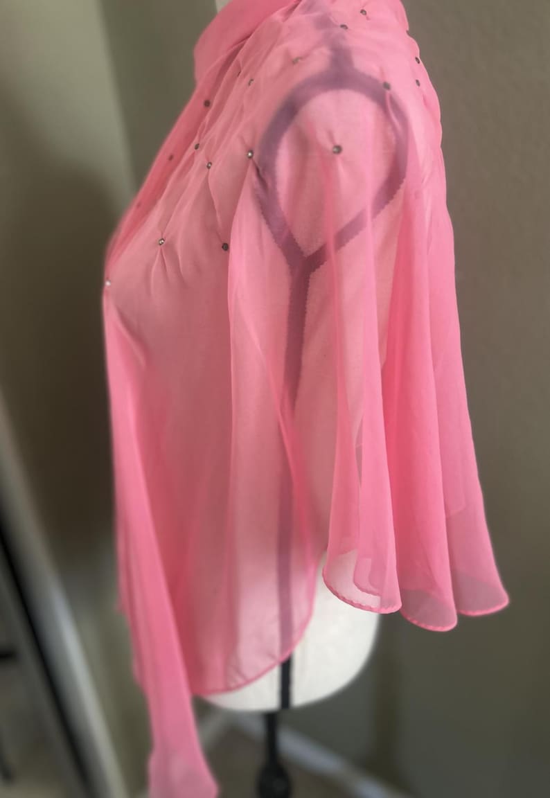 May include: A sheer, light pink top with a high collar and flared sleeves. The fabric is embellished with small, scattered silver-colored rhinestones. The garment is displayed on a white mannequin.
