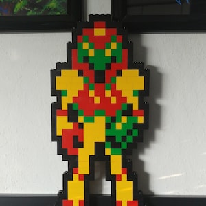 May include: A pixelated Lego figure of a video game character, Samus Aran from the Metroid series. The figure is made of red, yellow, green, and black Lego bricks.