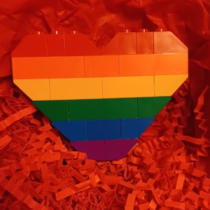 Può includere: Un cuore arcobaleno fatto di mattoncini Lego. Il cuore è fatto di mattoncini rossi, arancioni, gialli, verdi, blu e viola. Il cuore è seduto su un letto di carta velina rossa.