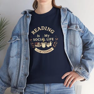 Op de afbeelding: Marineblauw T-shirt met de tekst "READING Is My SOCIAL LIFE CLUB FOUNDER" in een cirkelvormig ontwerp. Het ontwerp bevat boeken, een mok en een theepot. De persoon draagt een spijkerjas.