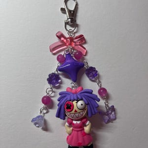 May include: A keychain featuring a doll with purple hair, a pink dress, and a button eye, suspended from a silver lobster clasp. The design includes a translucent pink bow, a large purple star bead, and dangling chains with purple flower and butterfly beads.
