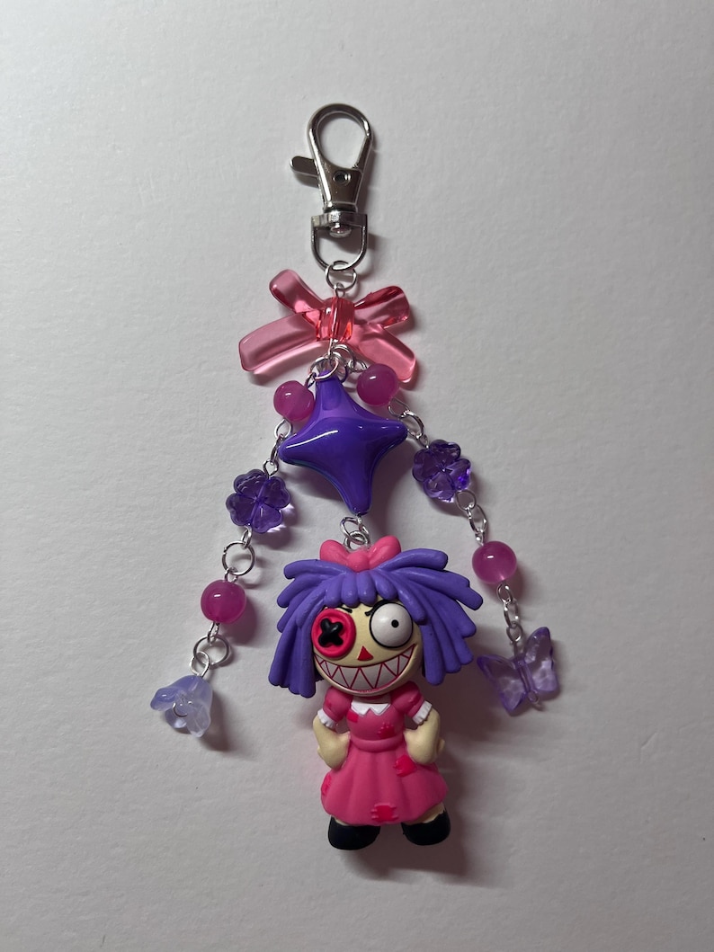 May include: A keychain featuring a doll with purple hair, a pink dress, and a button eye, suspended from a silver lobster clasp. The design includes a translucent pink bow, a large purple star bead, and dangling chains with purple flower and butterfly beads.