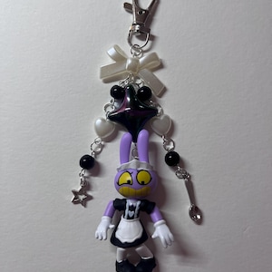 May include: A keychain featuring a purple cartoon rabbit figure wearing a black and white maid outfit. The charm is attached to a silver lobster clasp with a white ribbon bow, black and white beads, a dark star-shaped charm, and a small spoon.
