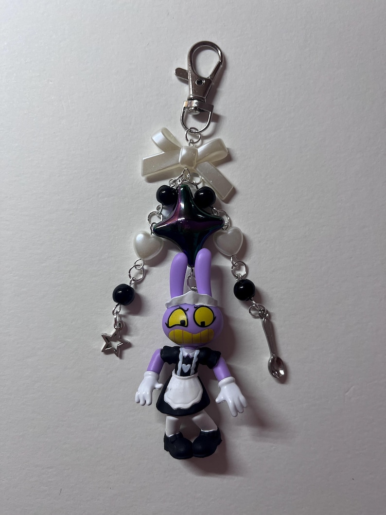 May include: A keychain featuring a purple cartoon rabbit figure wearing a black and white maid outfit. The charm is attached to a silver lobster clasp with a white ribbon bow, black and white beads, a dark star-shaped charm, and a small spoon.