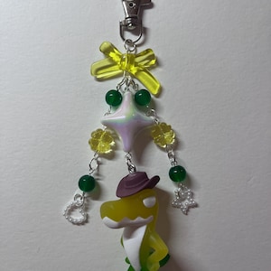 May include: A keychain featuring a yellow and green dinosaur figurine wearing a brown cowboy hat. The charm is decorated with a yellow translucent bow, a pearlescent star bead, green round beads, yellow flower beads, and small white heart and star charms.