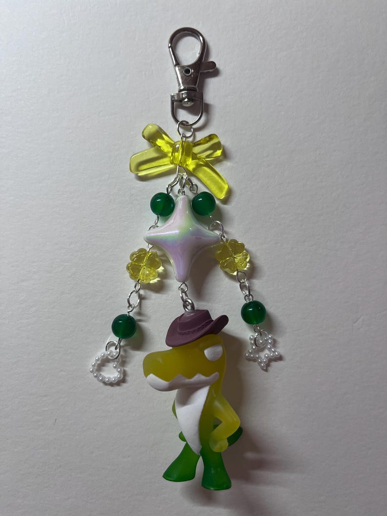 May include: A keychain featuring a yellow and green dinosaur figurine wearing a brown cowboy hat. The charm is decorated with a yellow translucent bow, a pearlescent star bead, green round beads, yellow flower beads, and small white heart and star charms.