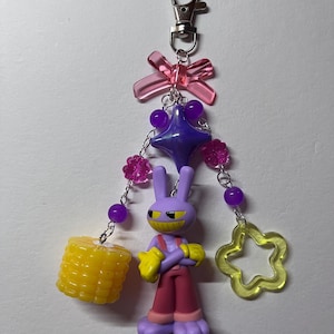 May include: A keychain featuring a purple cartoon rabbit figure with yellow gloves and red pants, accented with a translucent pink bow, a purple star bead, pink flower beads, a yellow corn charm, and a yellow open star charm on silver chains.