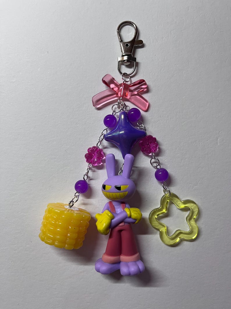 May include: A keychain featuring a purple cartoon rabbit figure with yellow gloves and red pants, accented with a translucent pink bow, a purple star bead, pink flower beads, a yellow corn charm, and a yellow open star charm on silver chains.