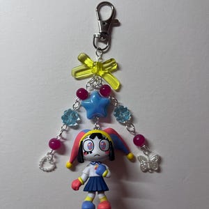 May include: A keychain featuring a cartoon jester figure with a blue and red hat, a white shirt, and a blue skirt. The charm is attached to a silver lobster clasp with a yellow bow, a blue star bead, light blue flower beads, pink beads, a white heart, and a butterfly.