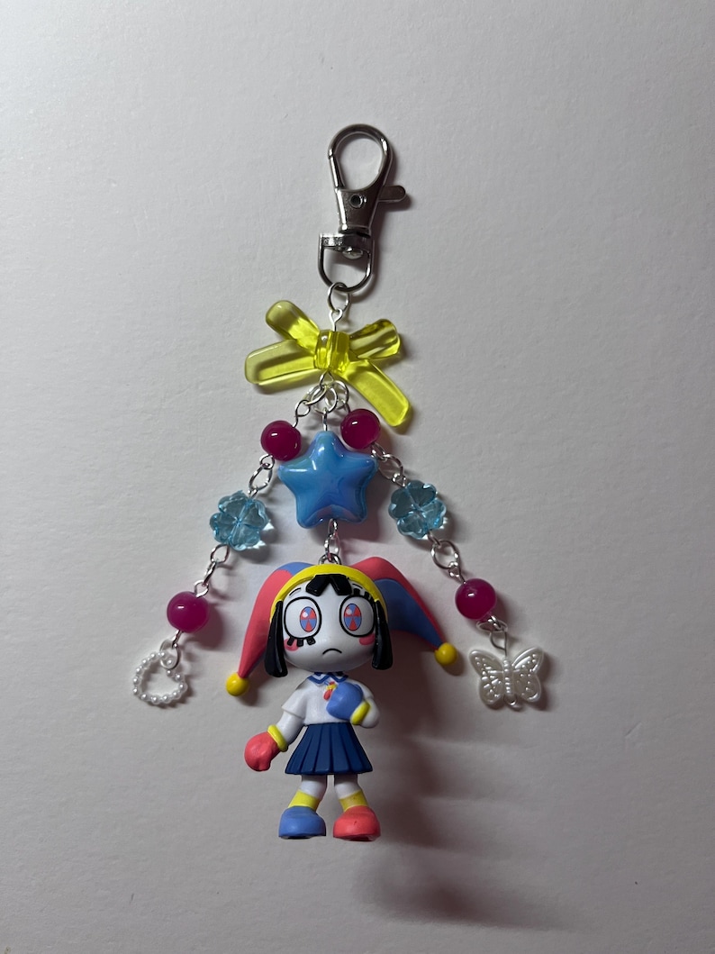 May include: A keychain featuring a cartoon jester figure with a blue and red hat, a white shirt, and a blue skirt. The charm is attached to a silver lobster clasp with a yellow bow, a blue star bead, light blue flower beads, pink beads, a white heart, and a butterfly.
