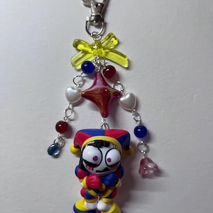 May include: A keychain featuring a jester figurine with a red, blue, and yellow outfit and hat. The charm is attached to a silver lobster clasp with a yellow translucent bow, a central star-shaped bead, and dangling strands of red, blue, and white heart beads.
