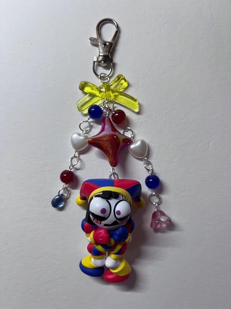 May include: A keychain featuring a jester figurine with a red, blue, and yellow outfit and hat. The charm is attached to a silver lobster clasp with a yellow translucent bow, a central star-shaped bead, and dangling strands of red, blue, and white heart beads.