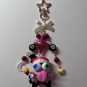 May include: A decorative keychain featuring a gold star clasp, a white bow, and a central character with a red mouth, white teeth, a pink tongue, and mismatched blue and green eyes. The figure wears a black top hat and holds a yellow microphone.