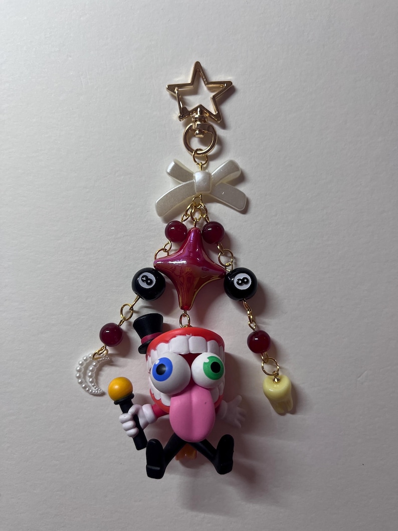 May include: A decorative keychain featuring a gold star clasp, a white bow, and a central character with a red mouth, white teeth, a pink tongue, and mismatched blue and green eyes. The figure wears a black top hat and holds a yellow microphone.