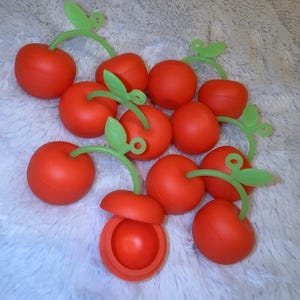 May include: A group of ten red cherry-shaped containers with green stems and leaves. One container is open, revealing a red interior.