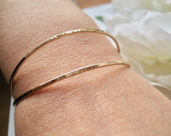 May include: Two gold hammered bangle bracelets stacked on a person's wrist. The bracelets are thin and delicate, with a hammered texture.