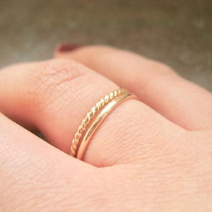 May include: Two gold rings on a finger. The top ring is a twisted band and the bottom ring is a simple band.