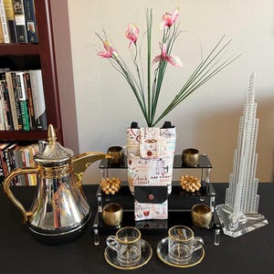 Op de afbeelding: Een zilveren en gouden koffiepot met een decoratief handvat, een set van vier kleine gouden kopjes en schoteltjes, een stoffen bekerhouder met de tekst "donkere branding" en "Slagroom toevoegen", een kralensnoer van hout en een glazen model van een wolkenkrabber.