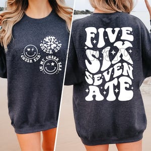 May include: A dark gray sweatshirt with white text and graphics. The front of the sweatshirt has three white circles with the text "Cheer Era" and a smiley face. The back of the sweatshirt has the text "Five Six Seven Ate" in a retro font.