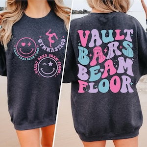 Puede incluir: Una sudadera gris oscura con un diseño gráfico rosa, azul y morado que dice "VAULT BARS BEAM FLOOR" en la espalda. La parte delantera de la sudadera tiene un diseño gráfico rosa y morado con una carita sonriente y las palabras "GYMNASTICS VAULT BARS BEAM FLOOR".