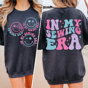 May include: A dark gray sweatshirt with a graphic design on the front and back. The front design features three smiley faces with the text "In My Sewing Era" around them. The back design features the text "In My Sewing Era" in large, colorful letters.