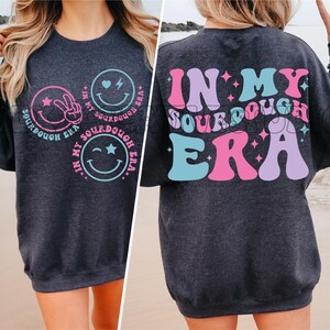 May include: A dark gray sweatshirt with the text "In My Sourdough Era" in pink, blue, and purple letters with stars. The front of the sweatshirt has three smiley faces with different expressions.