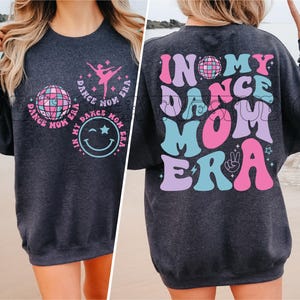 May include: A dark gray sweatshirt with a colorful graphic design on the front and back. The front design features a disco ball, a smiling face, and a ballerina. The back design features the text "In My Dance Mom Era" in a retro font.