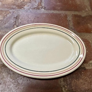 Op de afbeelding: Ovale, crèmekleurige serveerschaal met drie concentrische gekleurde banden: rood, groen en bruin. De rand van de schaal heeft een licht verhoogde rand. De schaal is gemaakt van keramiek en is geschikt voor het serveren van voedsel.