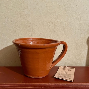 May include: A terracotta-colored ceramic mug with a wide, flared rim and a single handle. A rectangular paper tag is attached to the handle, featuring the text The Confederate Mug and Williamsburg Pottery Factory printed in black ink.