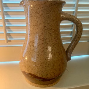 May include: A speckled beige ceramic pitcher with a brown glaze at the bottom. The pitcher has a wide mouth and a handle.