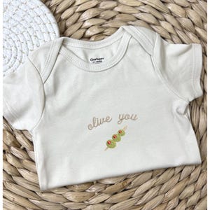 Puede incluir: Un body Gerber beige para bebé con mangas cortas. El texto "olive you" está bordado en cursiva, acompañado de un diseño de brocheta de aceitunas bordado. El body está colocado sobre una superficie tejida de tonos naturales.