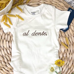 Puede incluir: Body de bebé color crema con el texto bordado "al dente" en marrón. El body está rodeado de pasta penne y flores sobre una esterilla tejida. El body es de la marca Gerber.