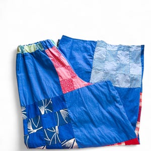 May include: A patchwork skirt in shades of blue, pink, and red. The skirt features a variety of fabric patterns, including floral, gingham, and solid colors. The waistband is elasticized and has a green and pink trim.