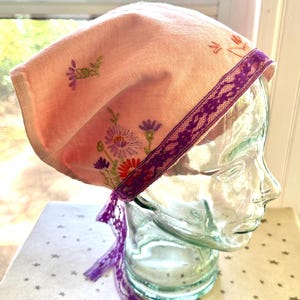 Puede incluir: Un cubrecabezas rosa claro con diseños florales bordados en morado, naranja y verde. El borde está adornado con una cinta de encaje morada y tiene cintas moradas a juego que cuelgan. El cubrecabezas se muestra en una cabeza de cristal.