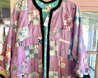 Puede incluir: Una chaqueta de patchwork con una mezcla de cuadrados de tela en color lavanda, estampados florales y cuadros. La prenda tiene un borde negro a lo largo de la parte delantera abierta y los puños, con un forro interior turquesa con lunares blancos y una etiqueta de la marca JN.