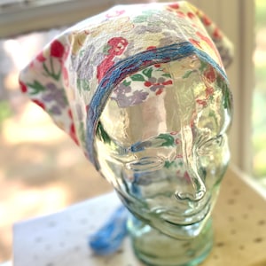 Könnte beinhalten: Ein Kopftuch mit Blumenmuster mit roten, lila und gelben Blumen auf weißem Grund. Das Kopftuch hat eine blaue Bordüre und wird auf einem Glaskopf präsentiert. Es handelt sich wahrscheinlich um einen Vintage- oder handgefertigten Artikel.