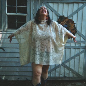 May include: A cream-colored lace poncho with a floral pattern. The person is wearing a white headband and black cowboy boots. The background is a light blue wooden shed with a window and a decorative piece.