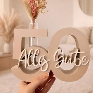 Può includere: Un oggetto decorativo in legno beige a forma di '50' con la scritta 'Alles Gute' in bianco. Contiene un piccolo vaso di vetro con fiori secchi. Il tutto è tenuto in mano.
