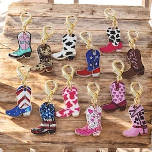 May include: A collection of gold-toned charm pendants in the shape of cowboy boots. The boots feature various designs, including floral patterns, cowhide prints, and patriotic themes. Each charm has a lobster clasp for easy attachment.