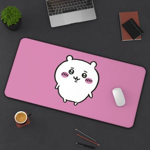 May include: A pink rectangular desk mat featuring a white cartoon character with a cute expression and pink cheeks. A white computer mouse is on the right. A laptop, notebook, and coffee cup are also visible.