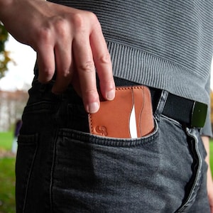 May include: A tan leather wallet with a white stripe is being put into a dark grey denim jeans pocket. The wallet has a curved design and a logo on the front. The person is wearing a grey ribbed jumper and a black belt.