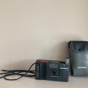 Puede incluir: Una cámara Olympus Trip MD negra con un botón rojo y una correa negra. La cámara está en una funda de cámara Olympus gris.