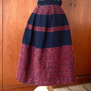 May include: A vintage midi skirt with a high waist. The skirt features a navy blue and red textured fabric. The skirt is displayed on a wooden dress form. The skirt has a gathered waistband.