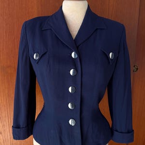 May include: A dark blue blazer featuring a notched lapel, three-quarter length sleeves with cuffs, and two decorative chest pockets. The front closure is secured with five circular buttons, each displaying a distinct white and dark blue pattern.